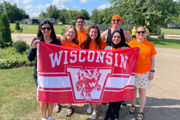 A group of seven people wearing orange t-shirts hold a large flag that says, "Wisconsin" with a "W" and Bucky Badger. They are in an outdoor setting, such as a campground.