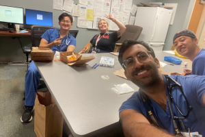 Four people in medical scrubs sitting at a table in a break room with food containers and supplies, smiling at the camera.
