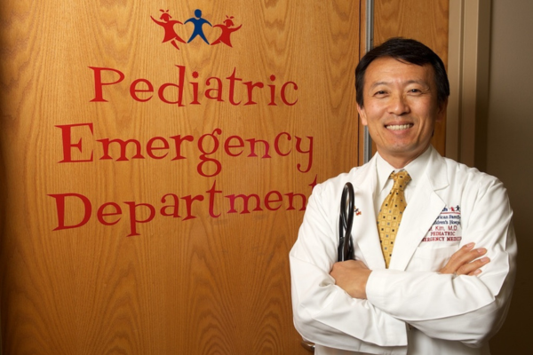 A pediatric emergency physician in a white coat stands smiling with arms crossed in front of a door labeled “Pediatric Emergency Department."