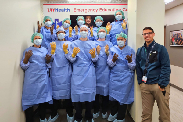 Group of trainees in blue medical gowns and gloves posing under a UW Health Emergency Education Center sign, with an instructor standing to the side.