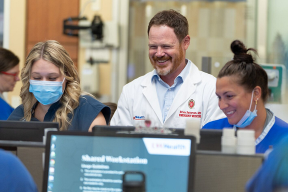 Medical staff gather at a shared workstation with computer monitors in a clinical setting.