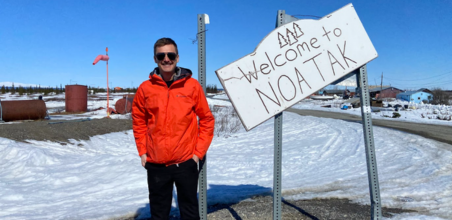 Dr. Mikko Sayre stands in front of a "Welcome to NOATAK" sign against a snowy background.