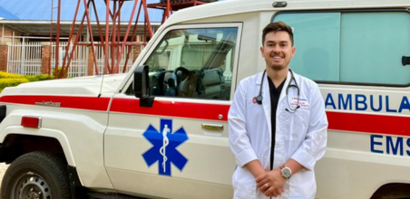 Person in a white coat with a stethoscope standing in front of an ambulance.