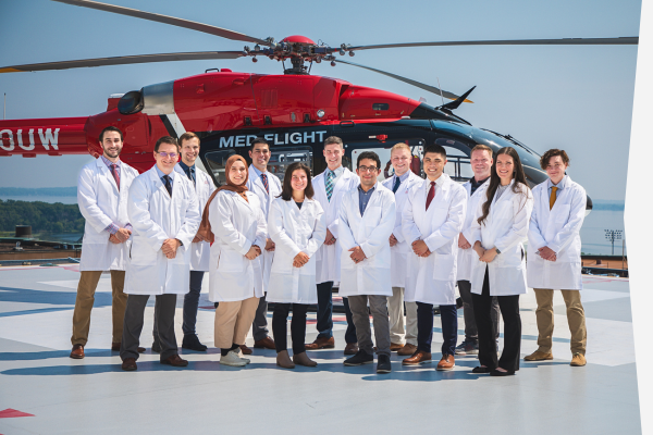 Group in white lab coats standing on a helipad, posing in front of a red medical helicopter with sky and water in the background.