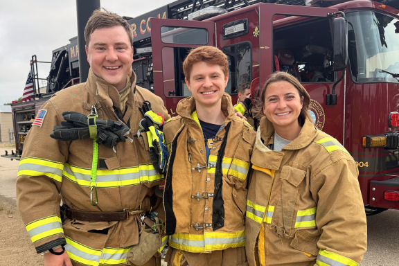Three residents pose together in front of a fire truck wearing firefighter gear.