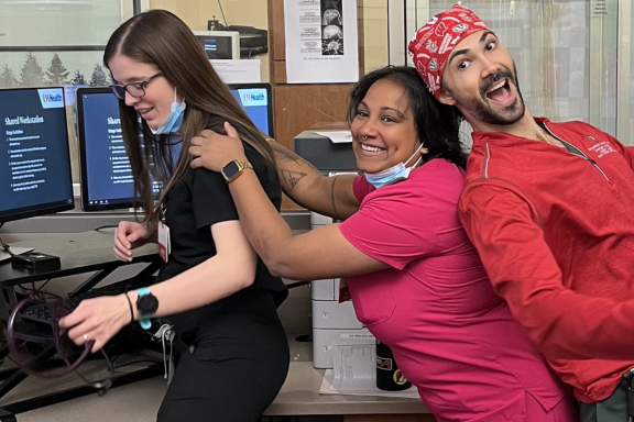 Three residents in scrubs stand back-to-back playfully near computer stations in a clinical office setting.