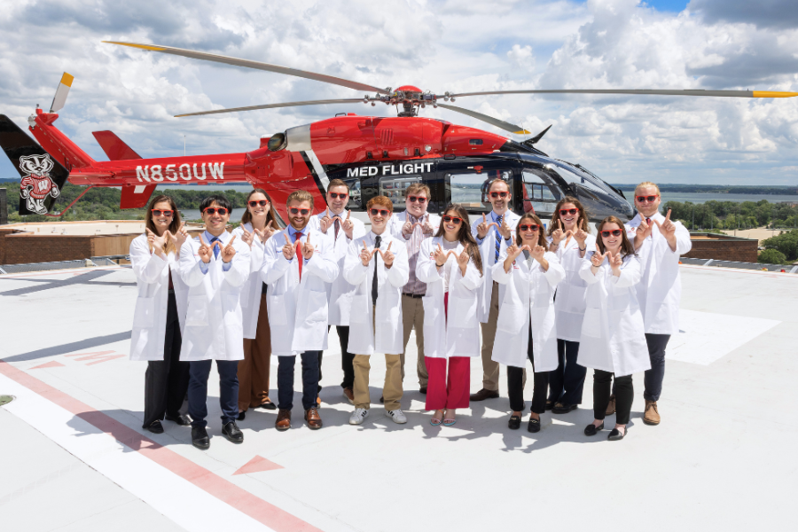 Group in white lab coats standing on a helipad, posing in front of a red medical helicopter with sky and water in the background.