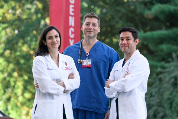 Three emergency medicine faculty pose in front of an Emergency sign wearing white coats and scrubs.